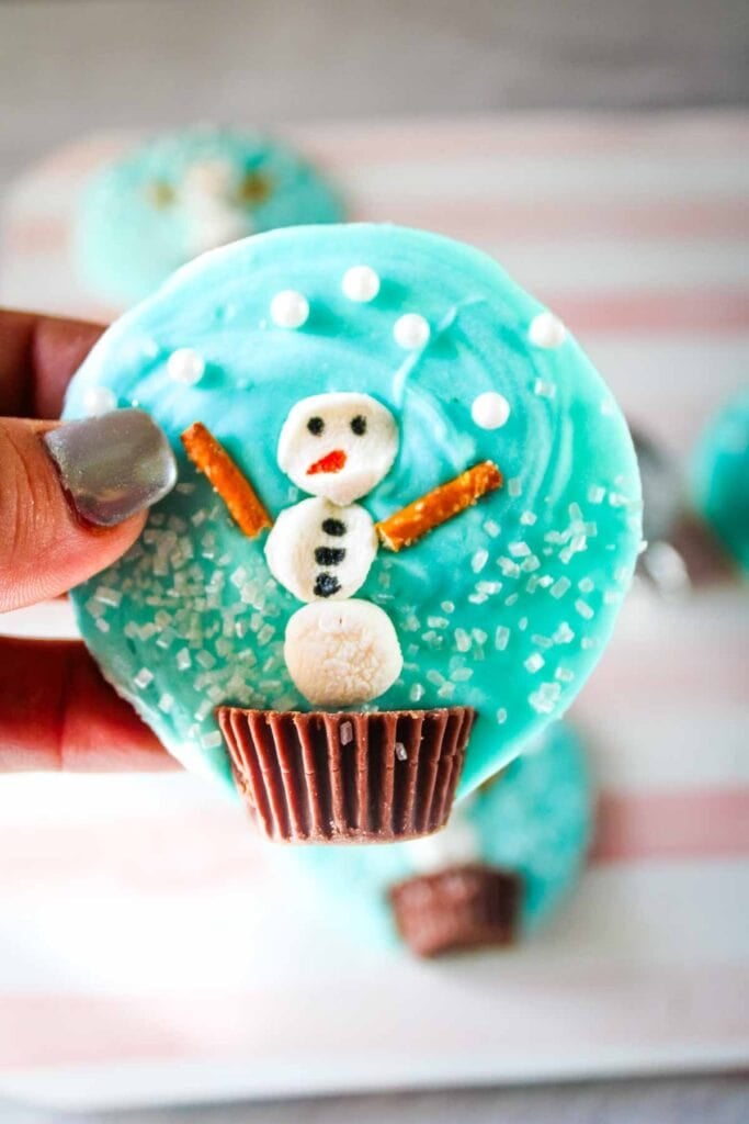 Blue-iced cookie topped with marshmallow snowman, pretzel arms, candy face, and peanut butter cup base, sprinkled with sugar crystals, held by hand with silver nails.