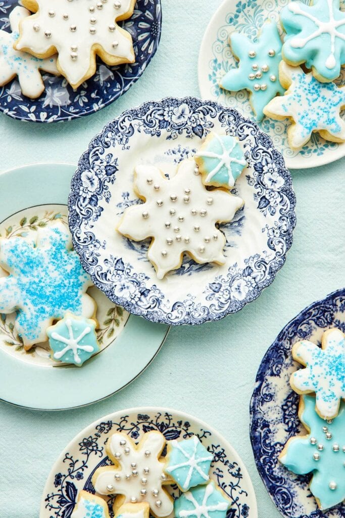 Snowflake-shaped cookies iced in white and light blue with sugar pearls and sprinkles, arranged on decorative blue-patterned plates.