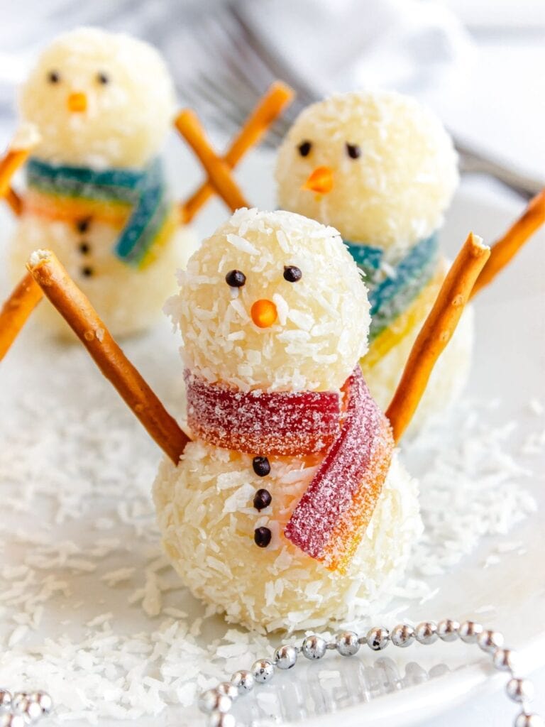 Coconut-coated snowman desserts with pretzel arms, candy faces, and gummy scarves, arranged on white plate sprinkled with coconut snow.