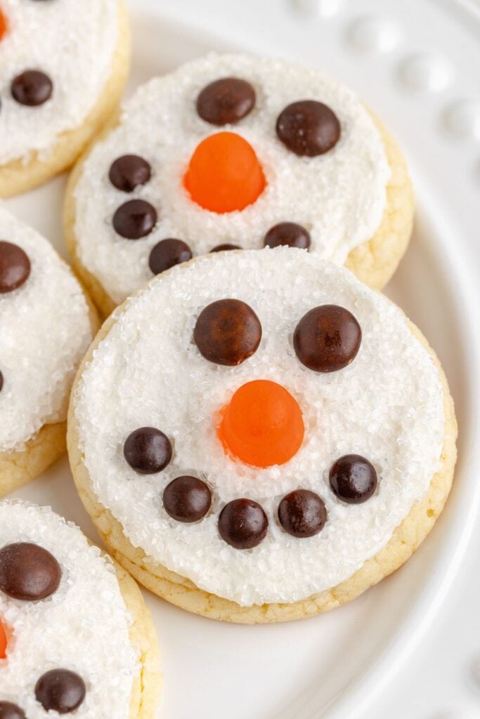 Round sugar cookies decorated as snowman faces with white icing, sugar crystal texture, candy eyes, orange nose, and chocolate smile, arranged on white plate.