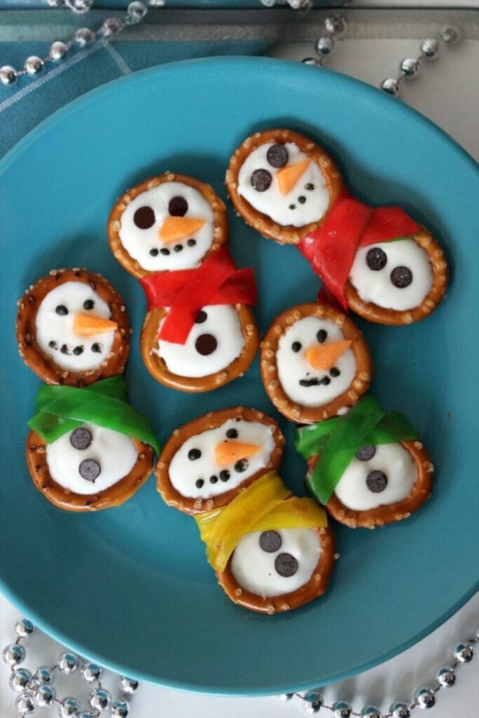 Pretzel snowmen with icing coating, chocolate chip faces, orange candy noses, and colorful candy scarves, arranged on blue plate with festive garland.