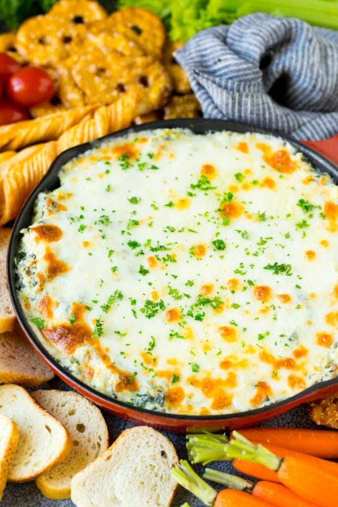 Skillet of baked spinach artichoke dip topped with golden cheese and parsley, surrounded by baguette slices, pretzels, breadsticks, tomatoes, celery, and carrots.