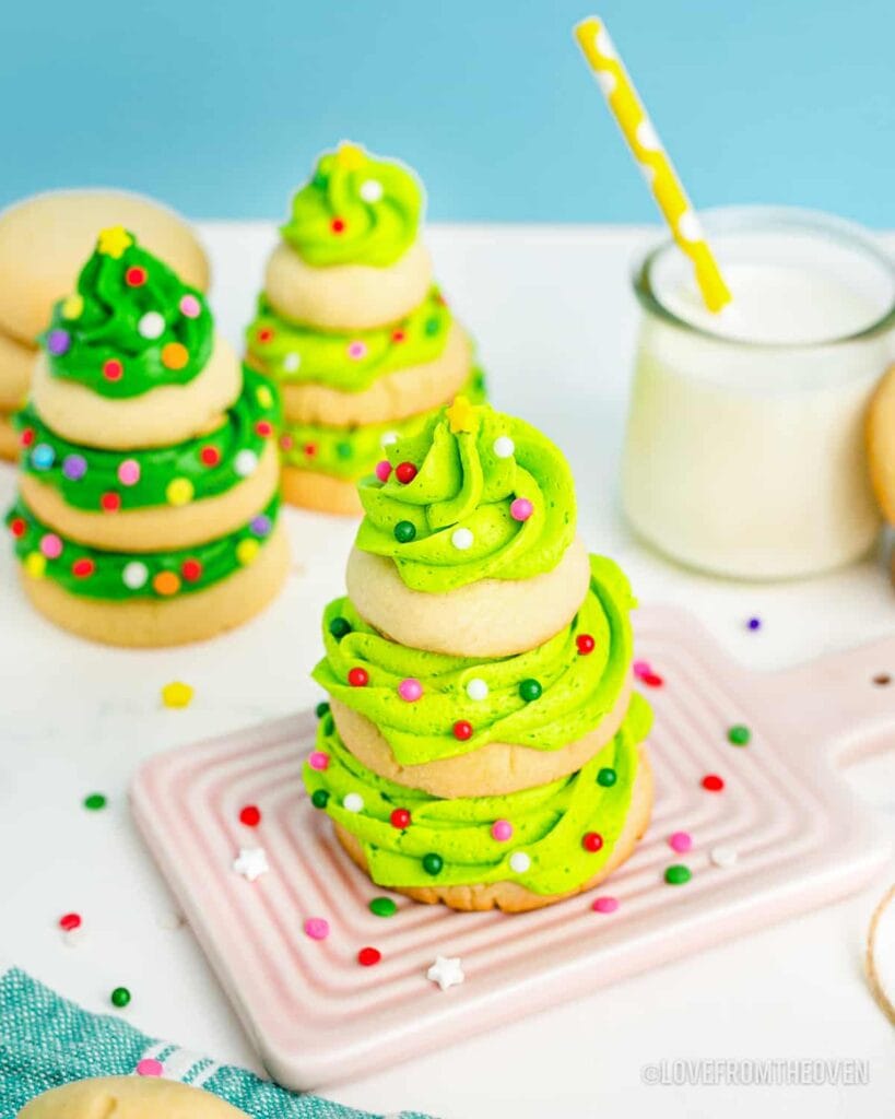 Stacked sugar cookies topped with swirled green frosting Christmas trees, decorated with sprinkles and yellow stars, arranged on pink tray with milk in background.
