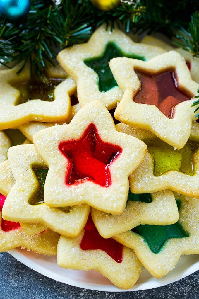 Star-shaped sugar cookies with colorful candy centers in red, green, and amber, arranged on plate near evergreen branches and ornaments.