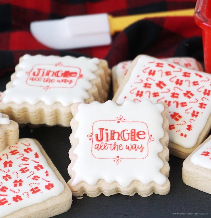 Square scalloped sugar cookies with white icing, decorated in red with “Jingle all the way” text, candy cane, and gift box designs, arranged on dark surface with plaid cloth and spatula.