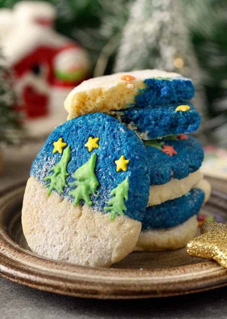 Round cookies with snowy icing and marshmallow snowmen heads, decorated with candy scarves, buttons, and stars, stacked on brown plate with powdered sugar and blurred holiday background.