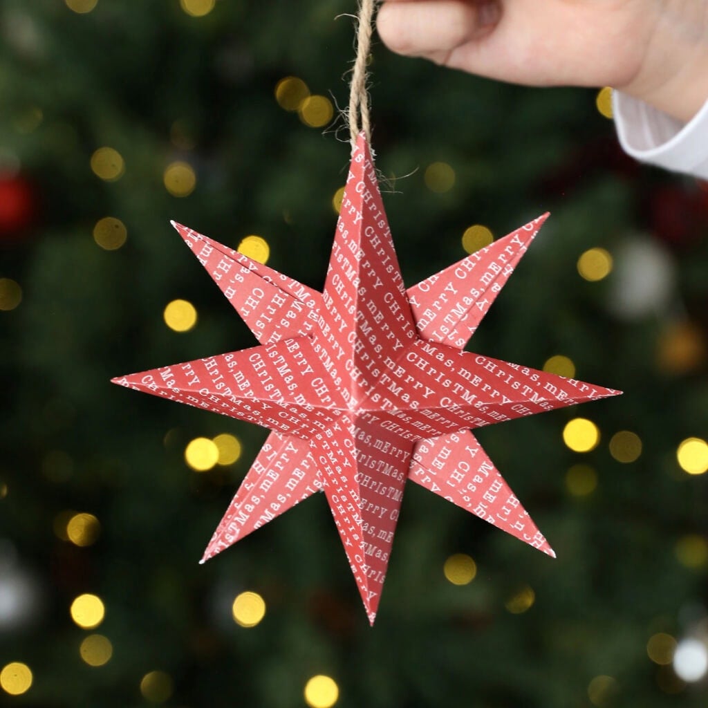 Red folded paper star ornament with “Merry Christmas” text, held by hand against blurred tree lights.
