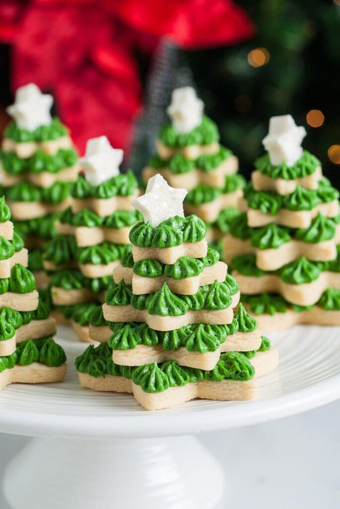 Stacked star-shaped sugar cookies with green frosting layers forming Christmas trees, topped with white stars, arranged on cake stand with festive blurred lights and poinsettias.