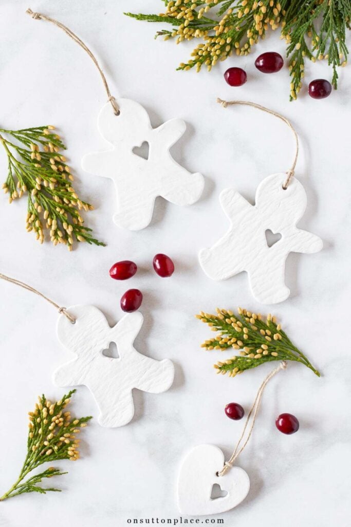 White clay ornaments shaped like gingerbread figures and heart with cutout centers, twine loops, arranged on marble surface with evergreen sprigs and cranberries.