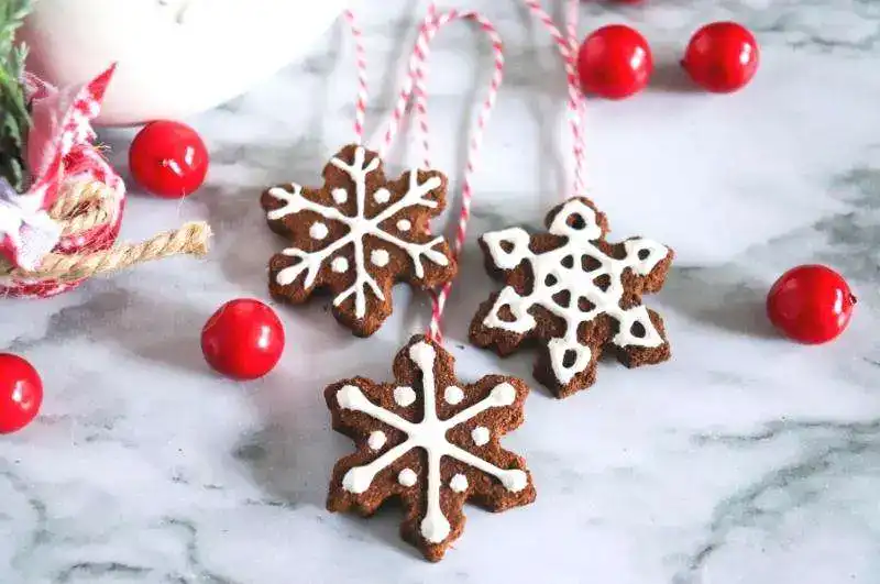 Three snowflake-shaped gingerbread cookies with white icing and red strings, arranged on marble surface with festive greenery and red decorations.