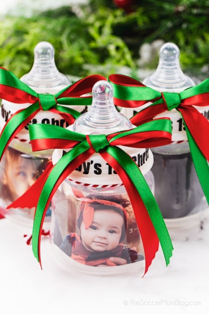 Three baby bottle ornaments with red and green bows, “Baby’s 1st Christmas” text, and baby photos inside, displayed with festive greenery.