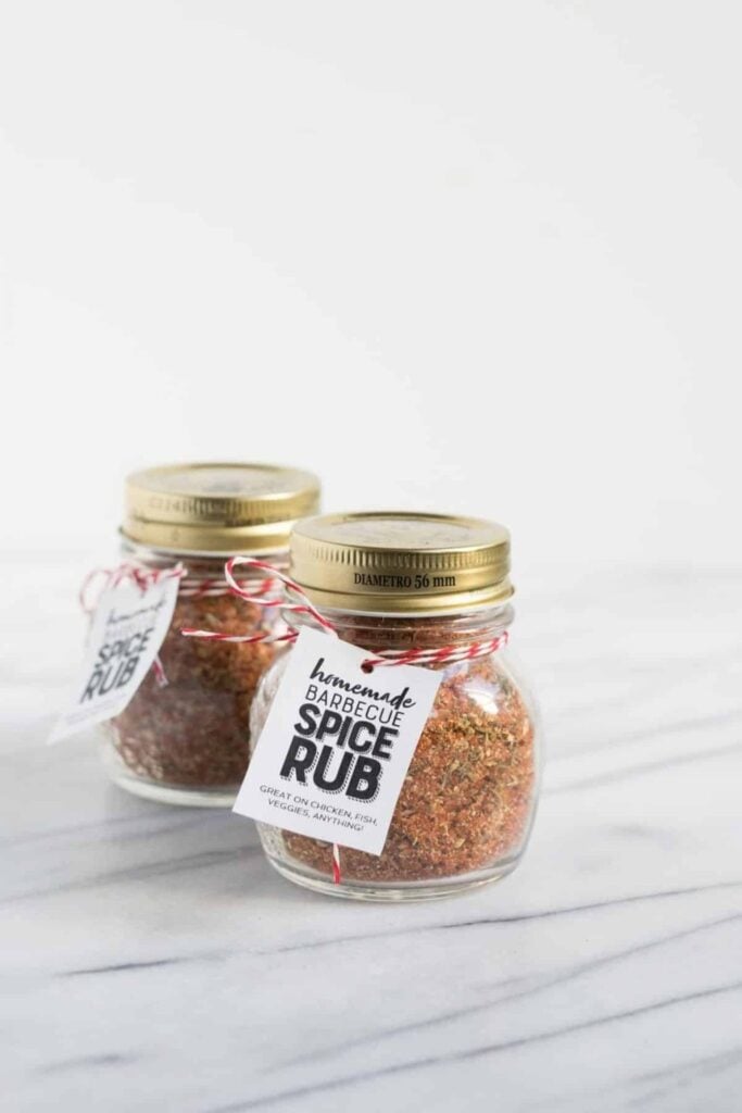 Two glass jars with gold lids, filled with reddish-brown barbecue spice rub, tied with red and white twine and tags reading “homemade BARBECUE SPICE RUB GREAT ON CHICKEN, FISH, VEGGIES, ANYTHING!”
