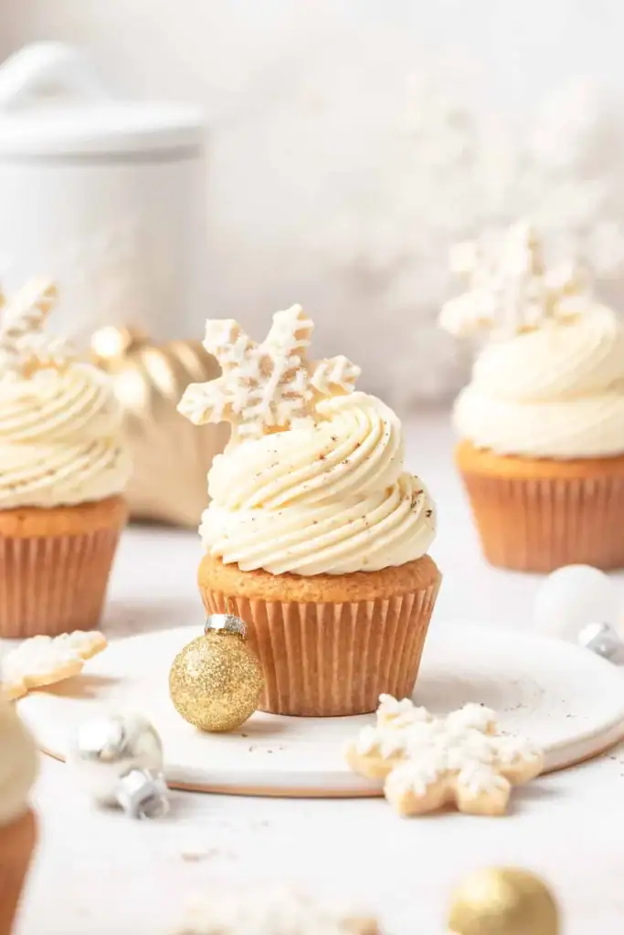 Cupcakes with white frosting swirls topped by snowflake cookies, dusted with sugar, arranged with gold and silver ornaments in soft winter setting.