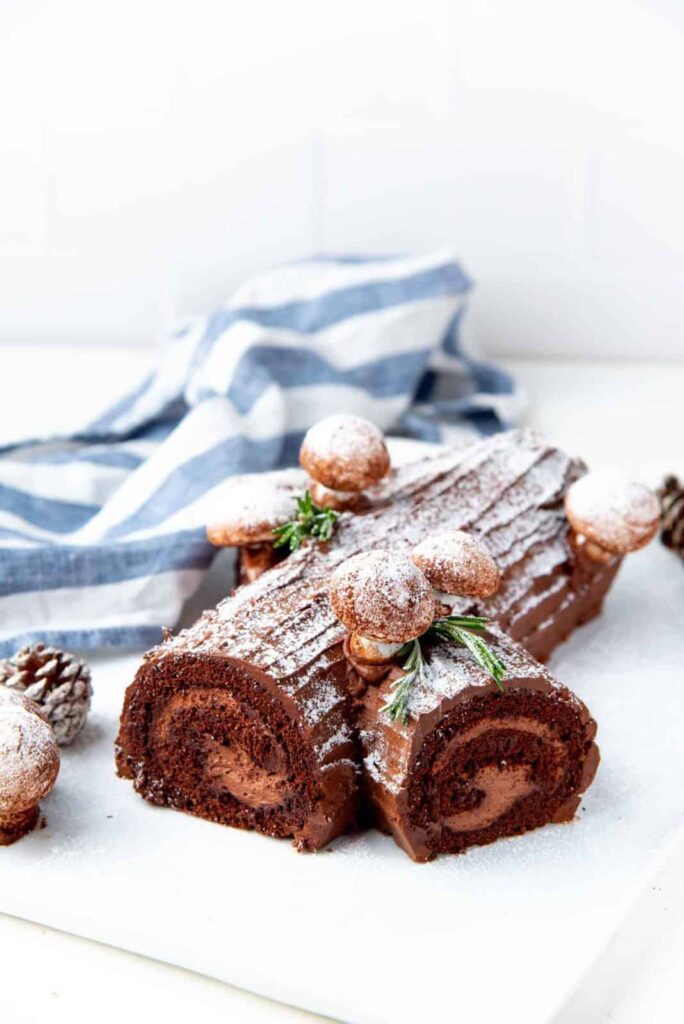 Chocolate Yule log cake with bark-like frosting, dusted with powdered sugar, topped with meringue mushrooms and greenery, displayed with pinecones and striped cloth in festive winter setting.
