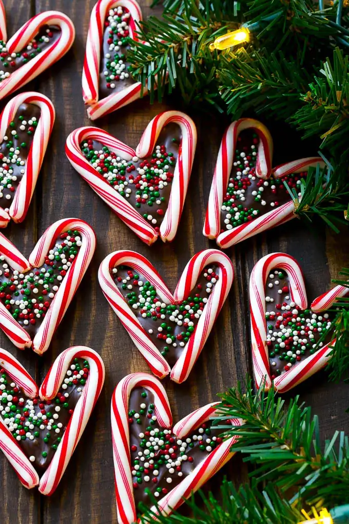 Heart-shaped candy canes filled with chocolate and topped with red, white, and green sprinkles, arranged on wooden surface with pine branches and Christmas lights.