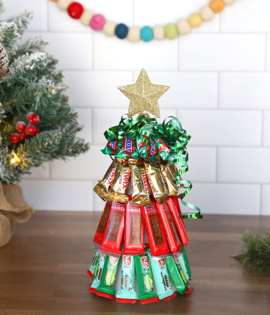 Christmas tree made of stacked chocolate bars topped with gold star and ribbon, set against tiled wall with garland and evergreen decor.