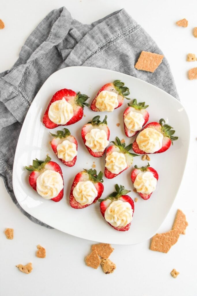 Halved strawberries filled with creamy topping and graham cracker crumbs, arranged on white plate with broken crackers and gray napkin nearby.