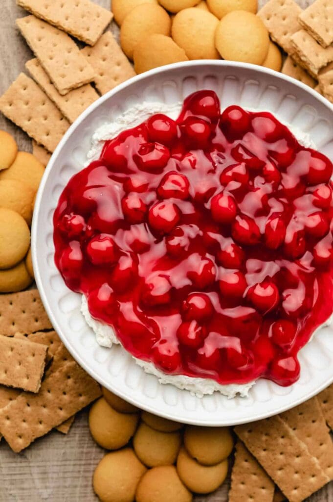White bowl of creamy dessert topped with glossy cherry pie filling, surrounded by graham crackers and vanilla wafers on wooden surface.