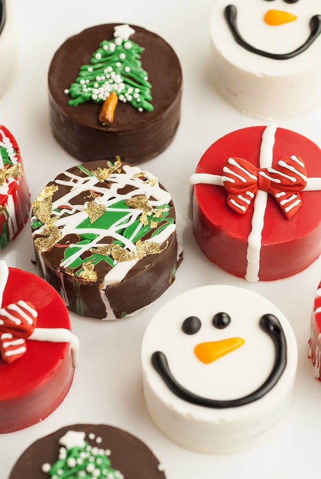 Assorted round holiday treats decorated as snowmen, gift boxes, and Christmas trees with colorful icing and gold accents, arranged in festive display.
