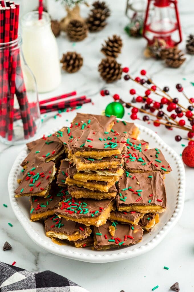 Stacked chocolate-covered toffee bark with red and green sprinkles, arranged on plate with pinecones, berries, ornament, lantern, and milk bottle with plaid straws.