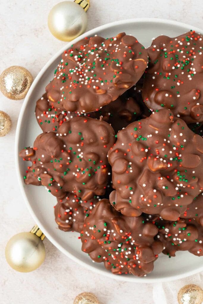 Chocolate nut clusters topped with red, green, and white sprinkles, arranged on white plate with gold and silver ornaments nearby.