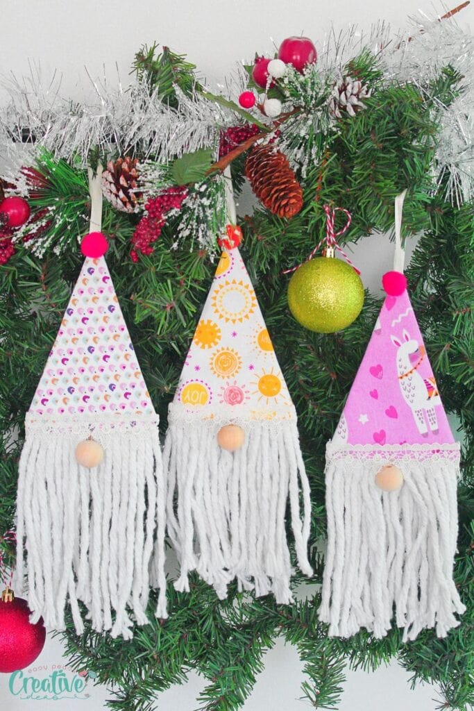 Three gnome ornaments with patterned hats, wooden bead noses, and yarn beards, hanging on decorated evergreen garland with pinecones, berries, tinsel, and ornaments.