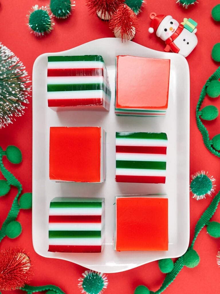 White plate of square red, white, and green layered gelatin desserts, surrounded by festive pom-poms, tinsel, and snowman ornament on red background.