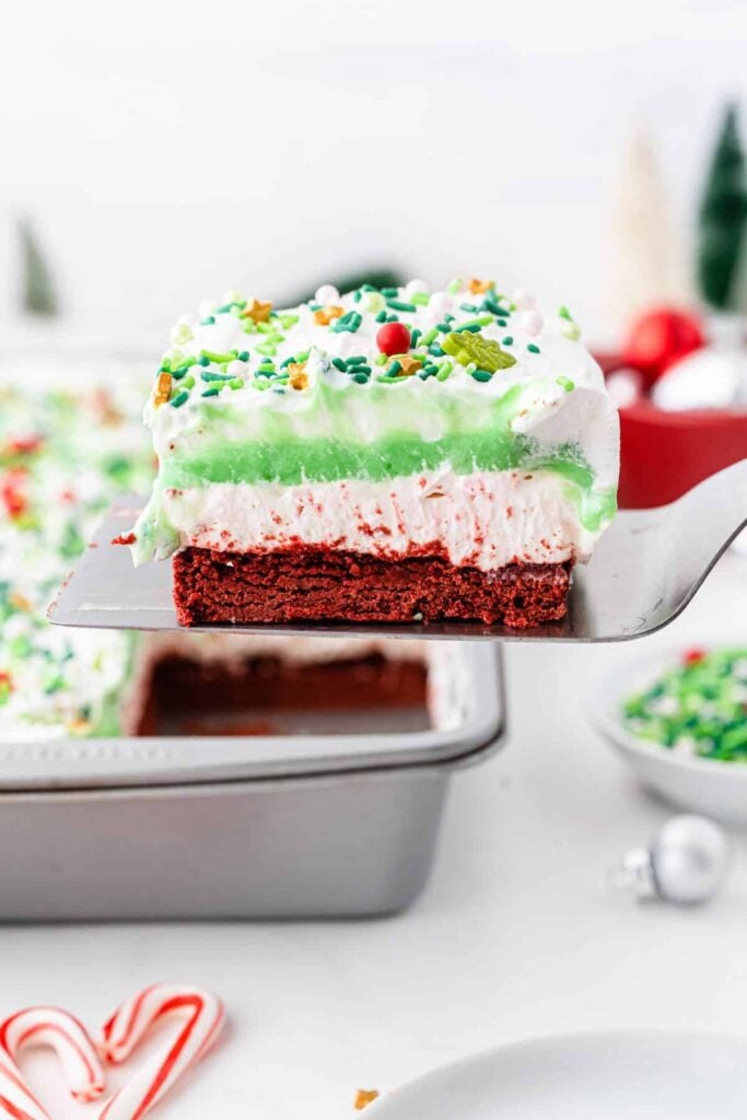 Red velvet base dessert bar layered with white cream and green pudding, topped with whipped cream and festive sprinkles, lifted from pan with blurred holiday decorations in background.