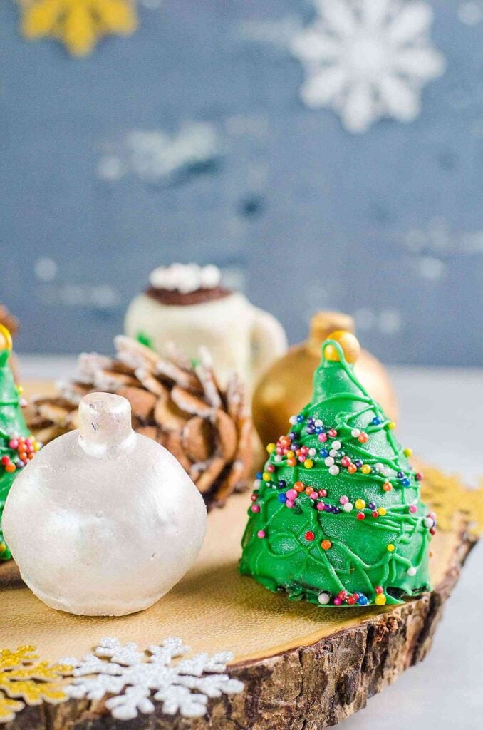 Festive green Christmas tree-shaped treat with sprinkles, displayed on wooden slab alongside ornament-like sweets, pinecones, and snowflake decorations in winter setting.