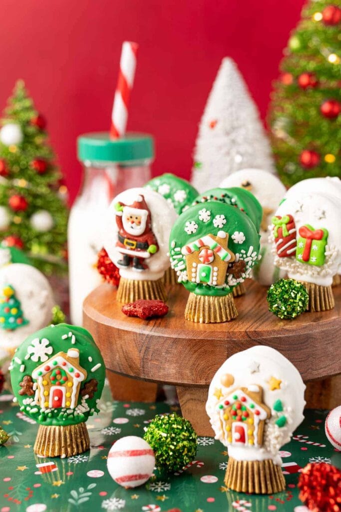 Christmas-themed treats decorated like snow globes with Santa, gingerbread houses, snowflakes, and presents, displayed on wooden stand with ornaments, trees, and milk bottle against red background.