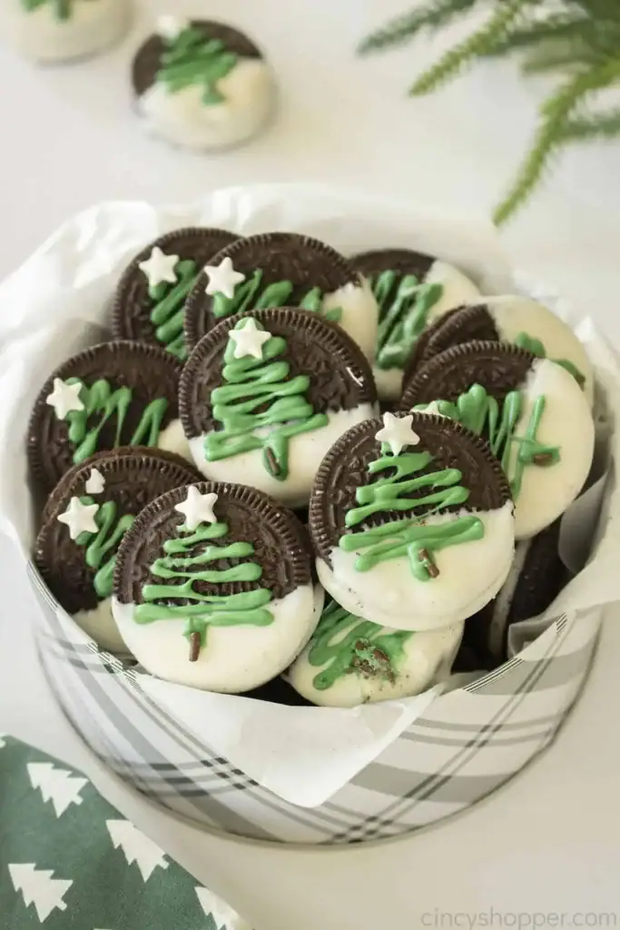 Chocolate sandwich cookies half-dipped in white chocolate, topped with green icing Christmas trees and white stars, arranged in plaid container with parchment lining and festive napkin nearby.