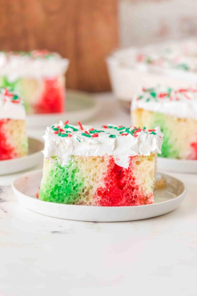 Slice of marbled red and green cake topped with white frosting and sprinkles, served on white plate with more slices in background.