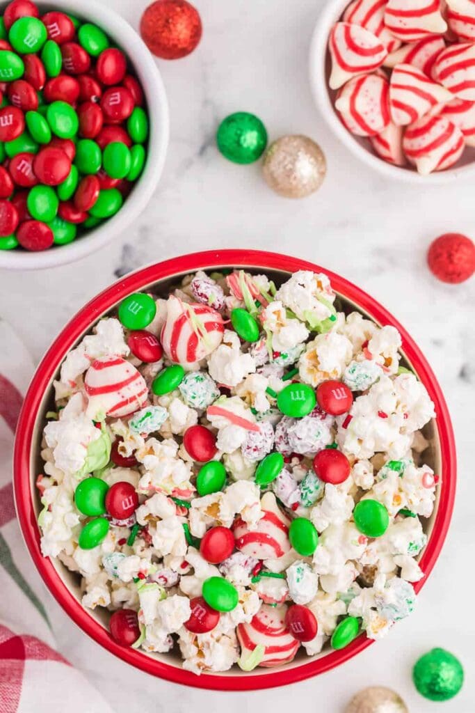 Red bowl of popcorn coated in white chocolate with red and green candies and peppermint pieces, surrounded by smaller bowls of M&Ms and peppermint, with glittery holiday decorations on marble surface.