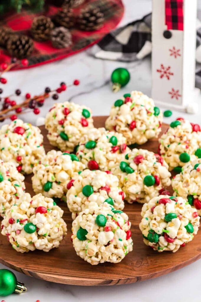 Wooden plate filled with round popcorn balls decorated with red and green candy pieces, surrounded by pinecones, plaid tray, ornaments, and snowflake décor in Christmas setting.