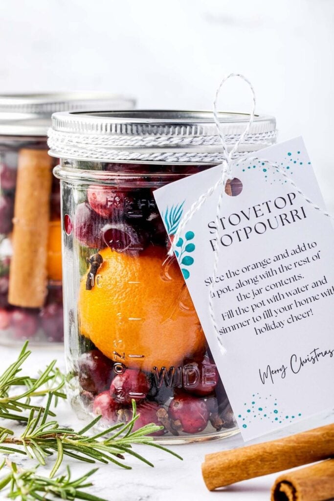 Glass jar filled with orange, cranberries, cloves, and cinnamon stick, tied with tag and string, surrounded by rosemary sprigs and cinnamon sticks on white surface.