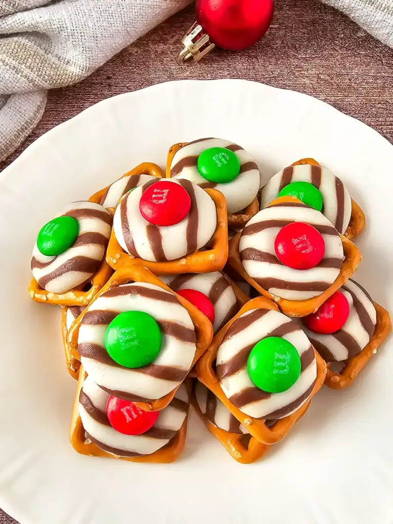 Square pretzels topped with striped chocolate and red or green candies, piled on white plate with festive background accents.