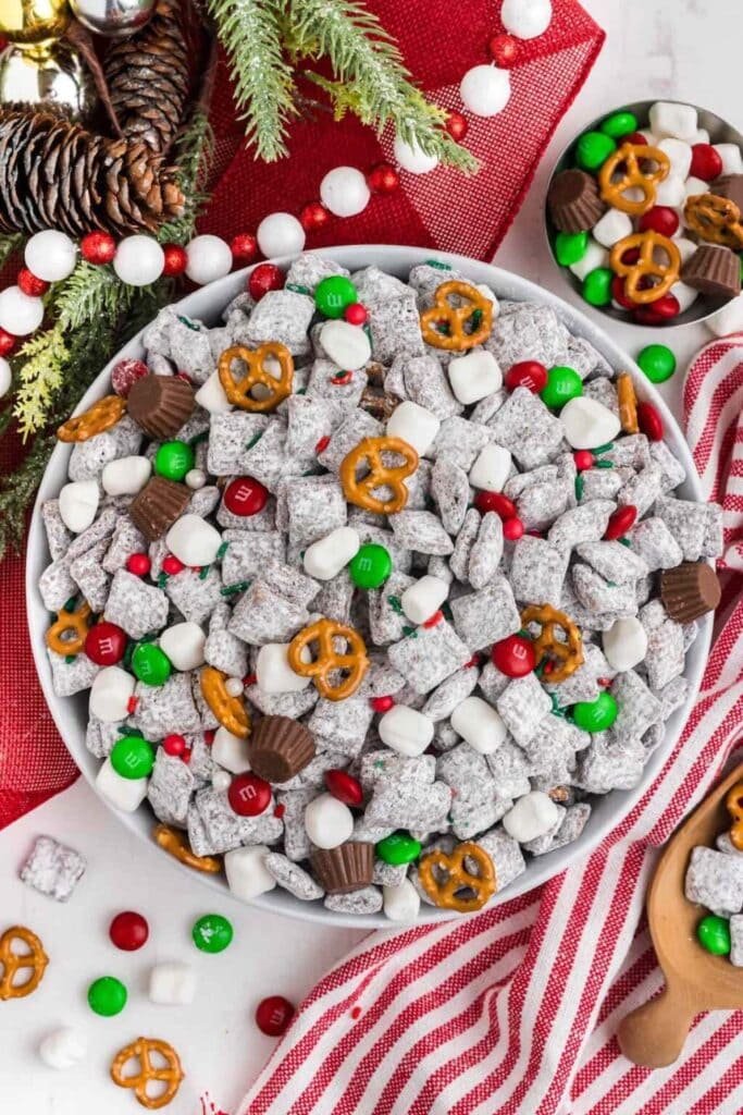 White bowl of festive snack mix with powdered cereal, pretzels, red and green candies, marshmallows, and peanut butter cups, surrounded by pinecones, evergreen, and red cloth.