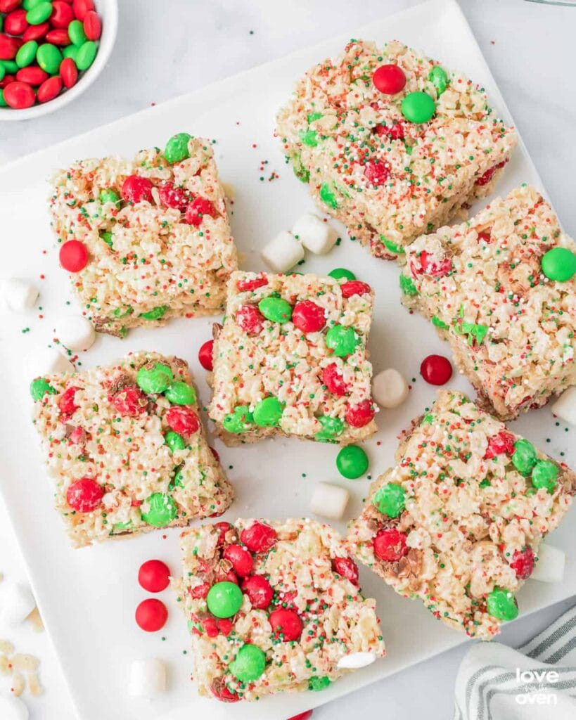 Square rice crispy treats topped with red and green candies and sprinkles, arranged on white plate with scattered marshmallows and small bowl of candies nearby.