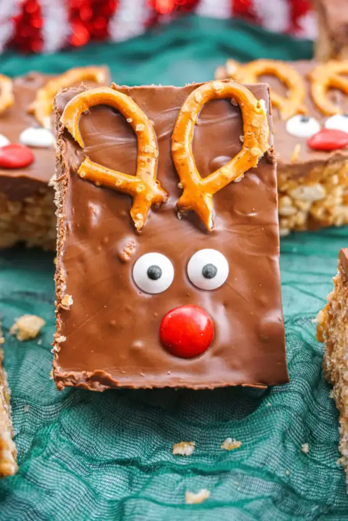 Chocolate-covered Rice Krispies treat decorated as reindeer with pretzel antlers, candy eyes, and red nose, placed on green cloth with similar treats in background.
