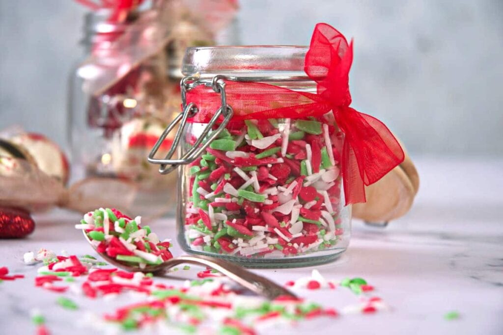 Glass jar of red, green, and white sprinkles tied with red ribbon, spoonful of sprinkles beside, scattered on marble surface with blurred holiday decor in background.