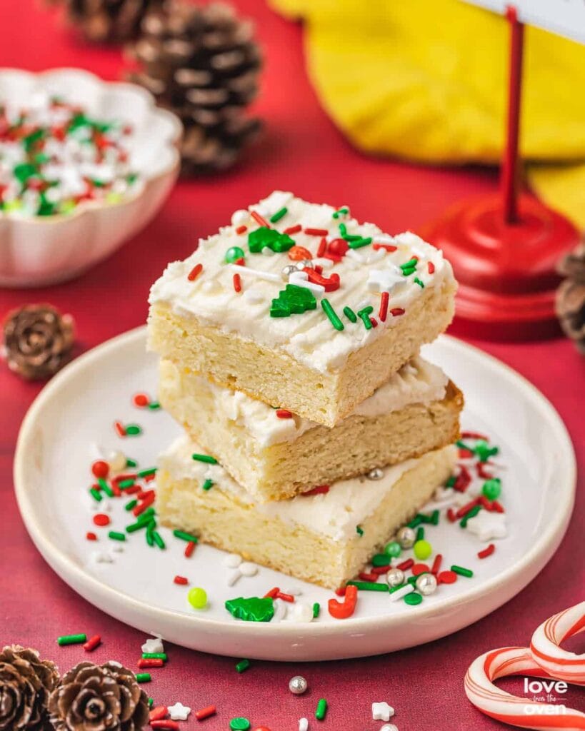 Stacked frosted sugar cookie bars topped with festive red, green, and white sprinkles, arranged on plate with scattered sprinkles, pinecones, and colorful background accents.