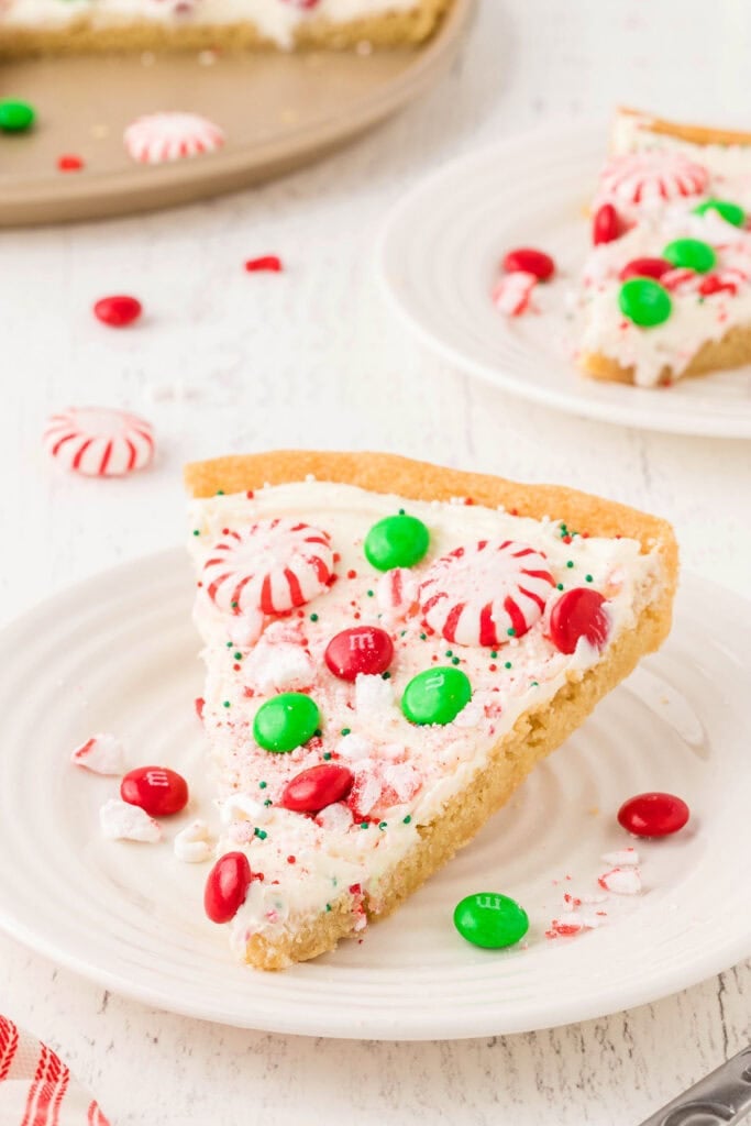Slice of cookie-crust dessert pizza topped with white frosting, peppermint candies, red and green chocolates, and crushed peppermint, served on white plate with more slices nearby.