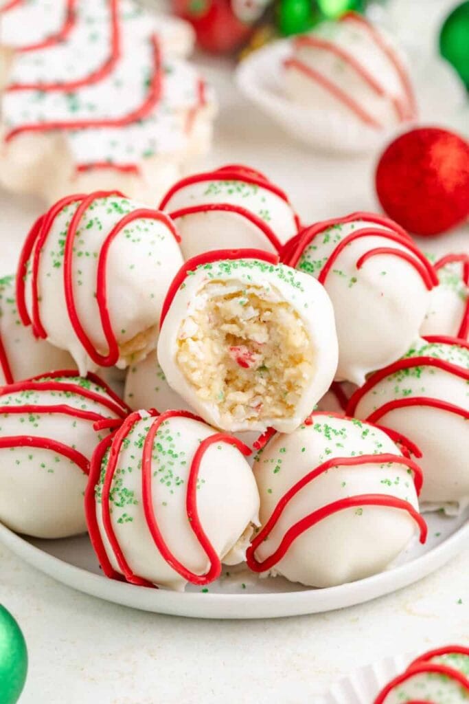 White chocolate-coated dessert balls with red icing drizzle and green sprinkles, one bitten to reveal creamy interior with colorful bits, arranged on plate with blurred ornaments in background.