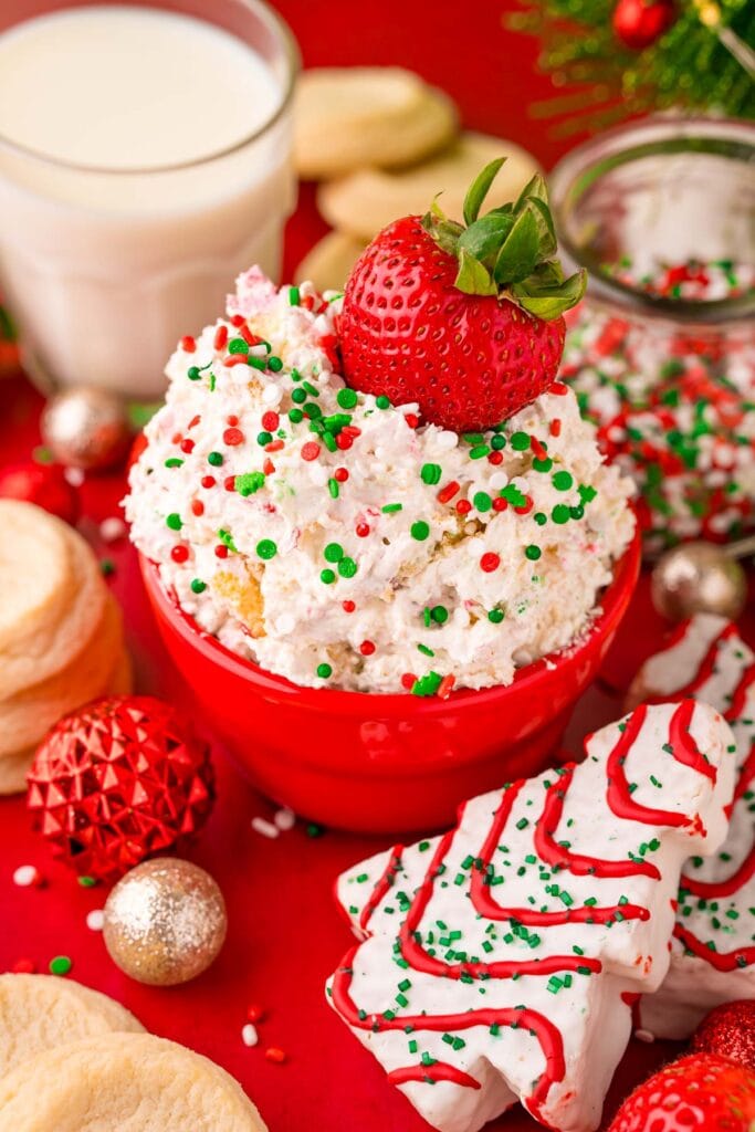 Red bowl of creamy dip topped with sprinkles and strawberry, surrounded by Christmas tree cookies, sugar cookies, ornaments, and glass of milk on red background.