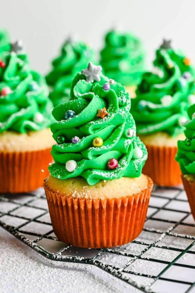Cupcakes with green frosting shaped like Christmas trees, decorated with colorful sprinkles and stars, lightly dusted with powdered sugar, arranged on cooling rack.