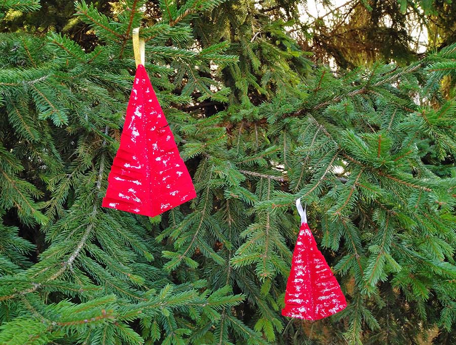 Two red triangular Christmas tree ornaments with snowy speckles, hanging by gold and white ribbons on pine branches.