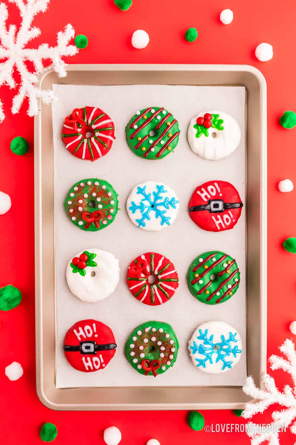 Ring-shaped cookies decorated with colorful icing patterns, festive designs like stripes, holly, snowflakes, and Santa belts, arranged on tray with red background and pom-poms.
