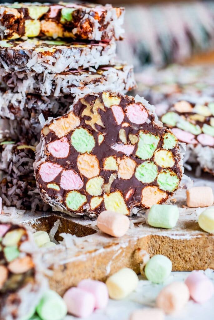Slices of chocolate fudge with multicolored marshmallows coated in coconut, stacked on wooden board with loose pastel marshmallows scattered around.