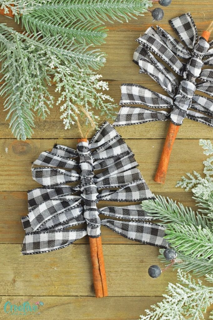 Two handmade Christmas tree ornaments from cinnamon sticks and checkered ribbon, arranged with snowy evergreen sprigs and berries on wooden surface.