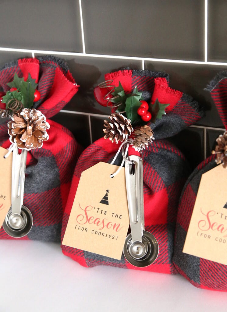 Three plaid fabric gift bags tied with pinecones, holly, and berries, each with tag reading “’TIS THE SEASON (FOR COOKIES)” and attached measuring spoon.